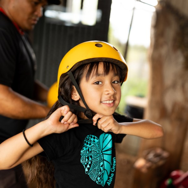 Child smiling, wearing a yellow helmet indoors with an adult nearby.