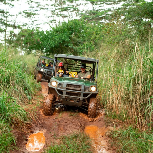 Two off-road vehicles drive through muddy, grassy terrain with passengers.