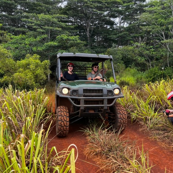Two people driving an off-road vehicle on a dirt path with tall grass, another person photographing nearby.