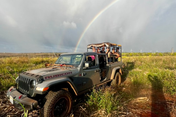 Jeep with people on a grassy field under a rainbow in a cloudy sky.