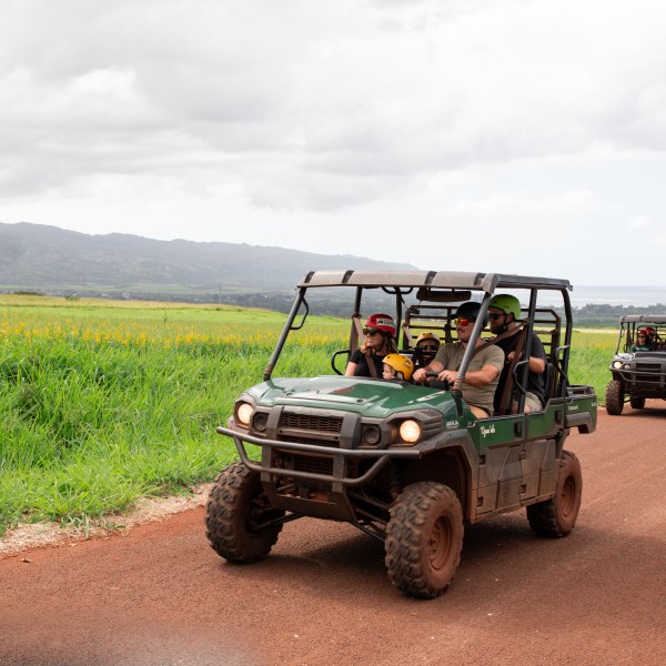 Two off-road vehicles driving on a dirt path through grassy fields with mountains in the background.