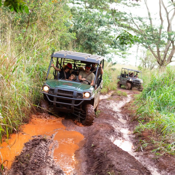 Two off-road vehicles drive through a muddy, forested trail.